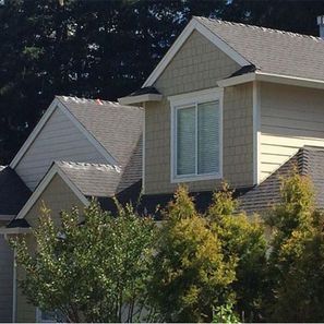 Beige house with multiple gables and a white framed window; green trees in the foreground.