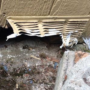 Damaged beige vent near a concrete foundation. Torn metal strips hang down; debris on the ground.