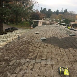 Roof with worn shingles being cleaned, with greenery and other buildings in the background.