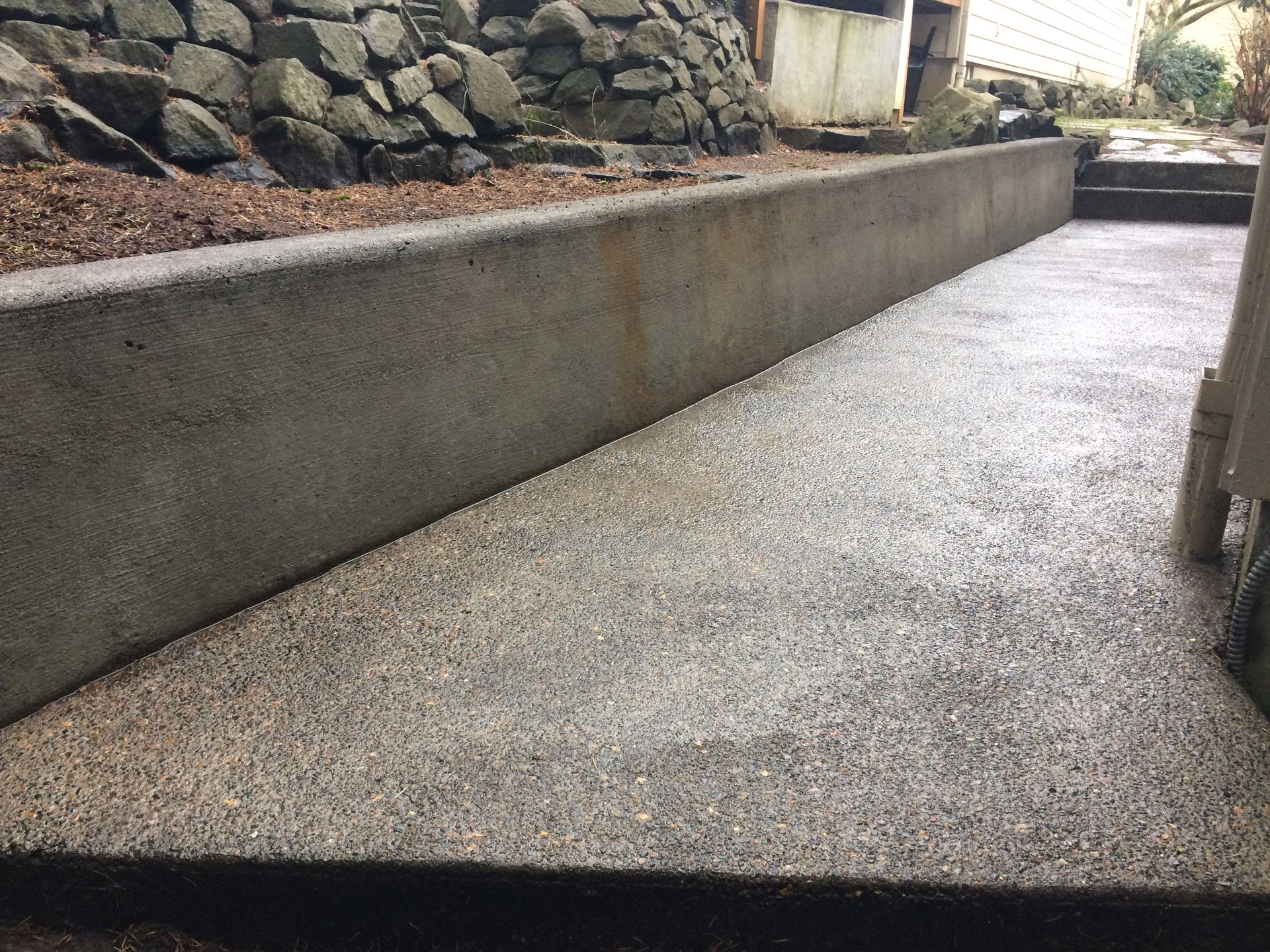 Concrete retaining wall next to pebbled pathway. Rocky ground at top, stairs visible on right.