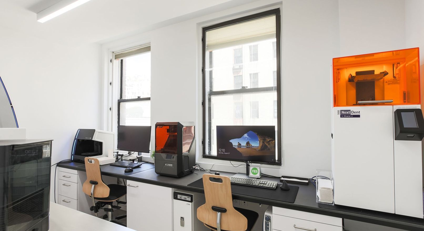 Laboratory with computers, 3D printer, and workstations; bright, white interior, windows, and two chairs.
