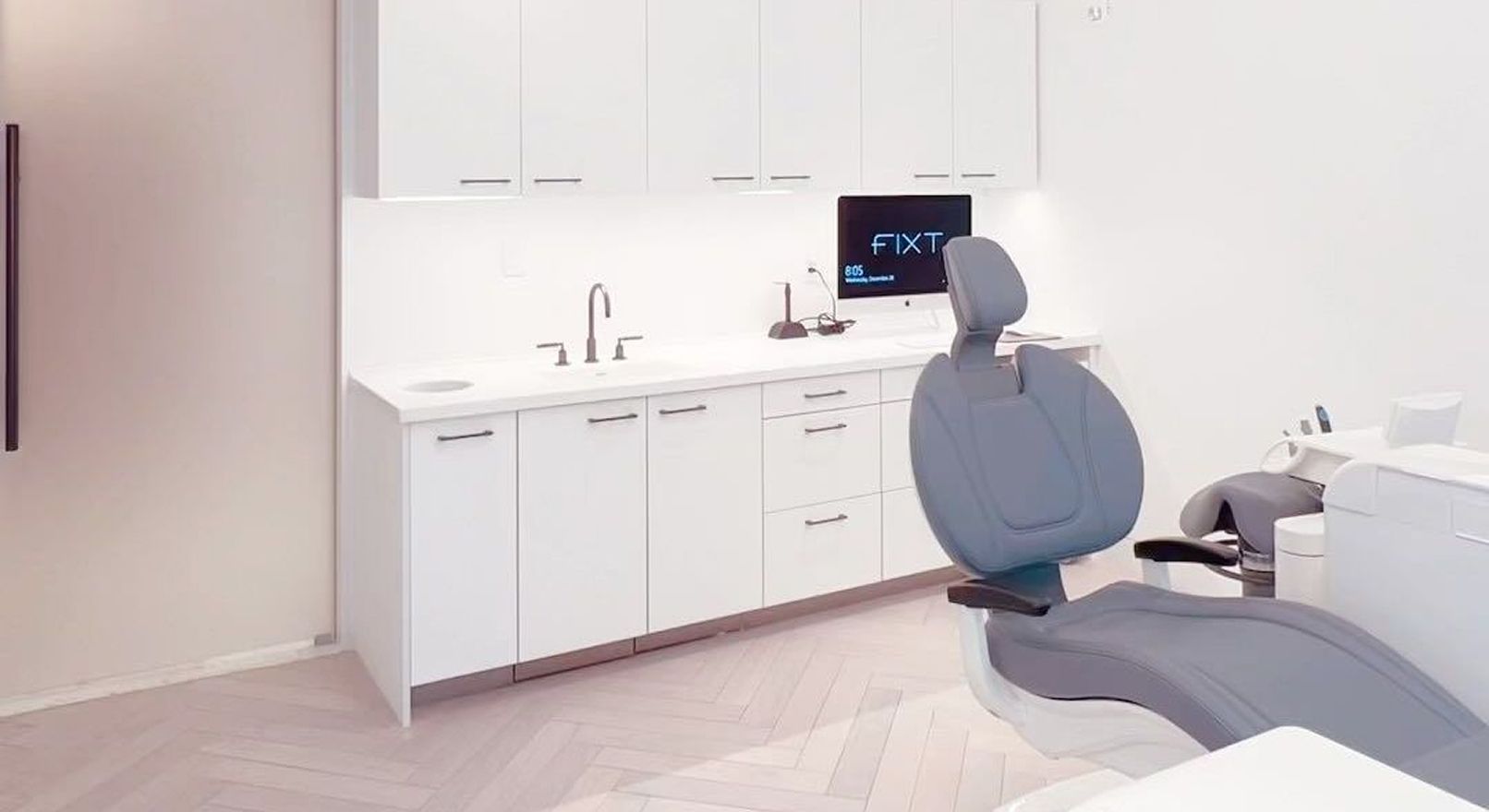 A dental office with a gray chair, white cabinets, and a sink.
