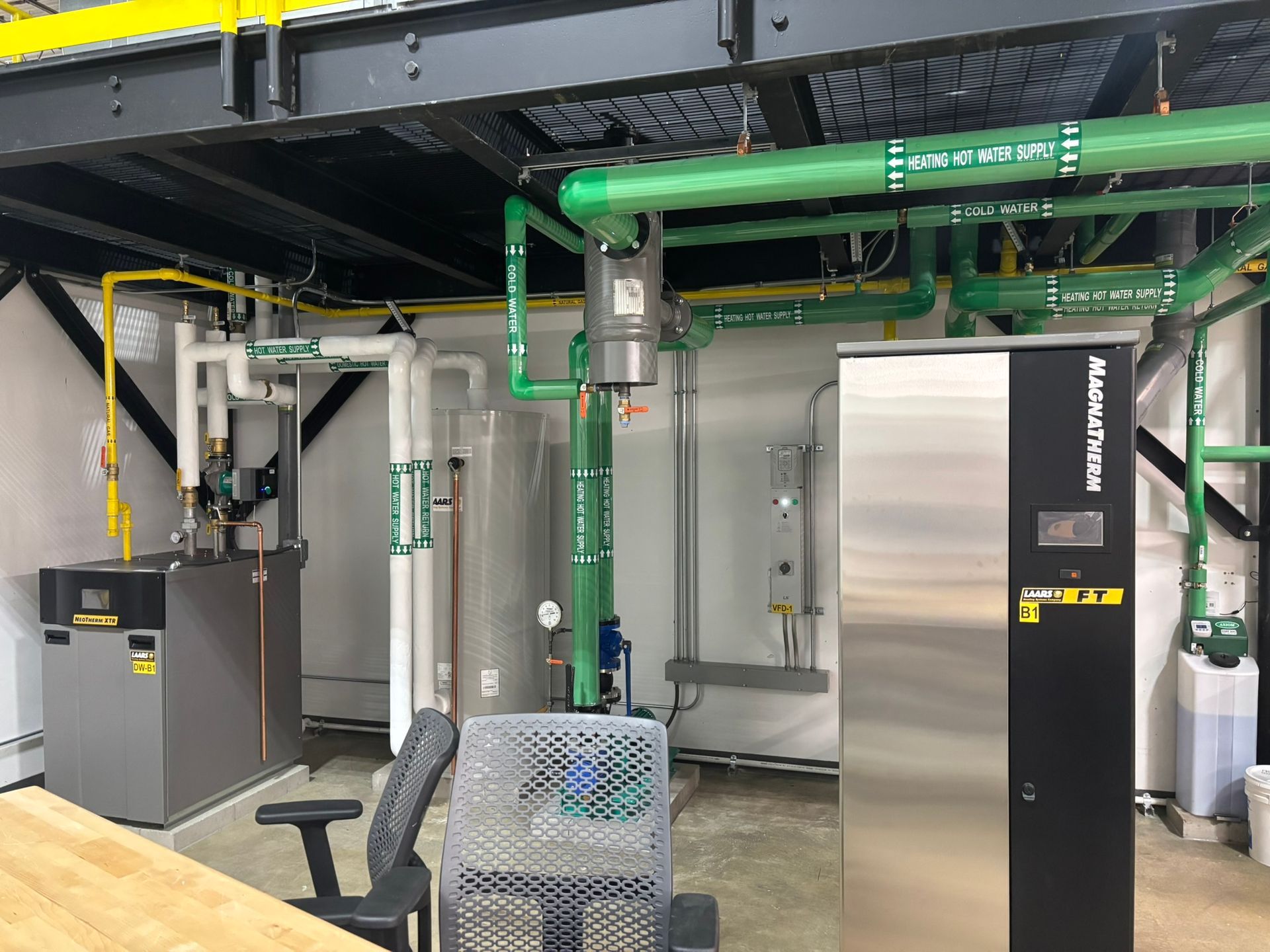 Mechanical room with boilers, pipes, and control panels. Green and white pipes are visible.