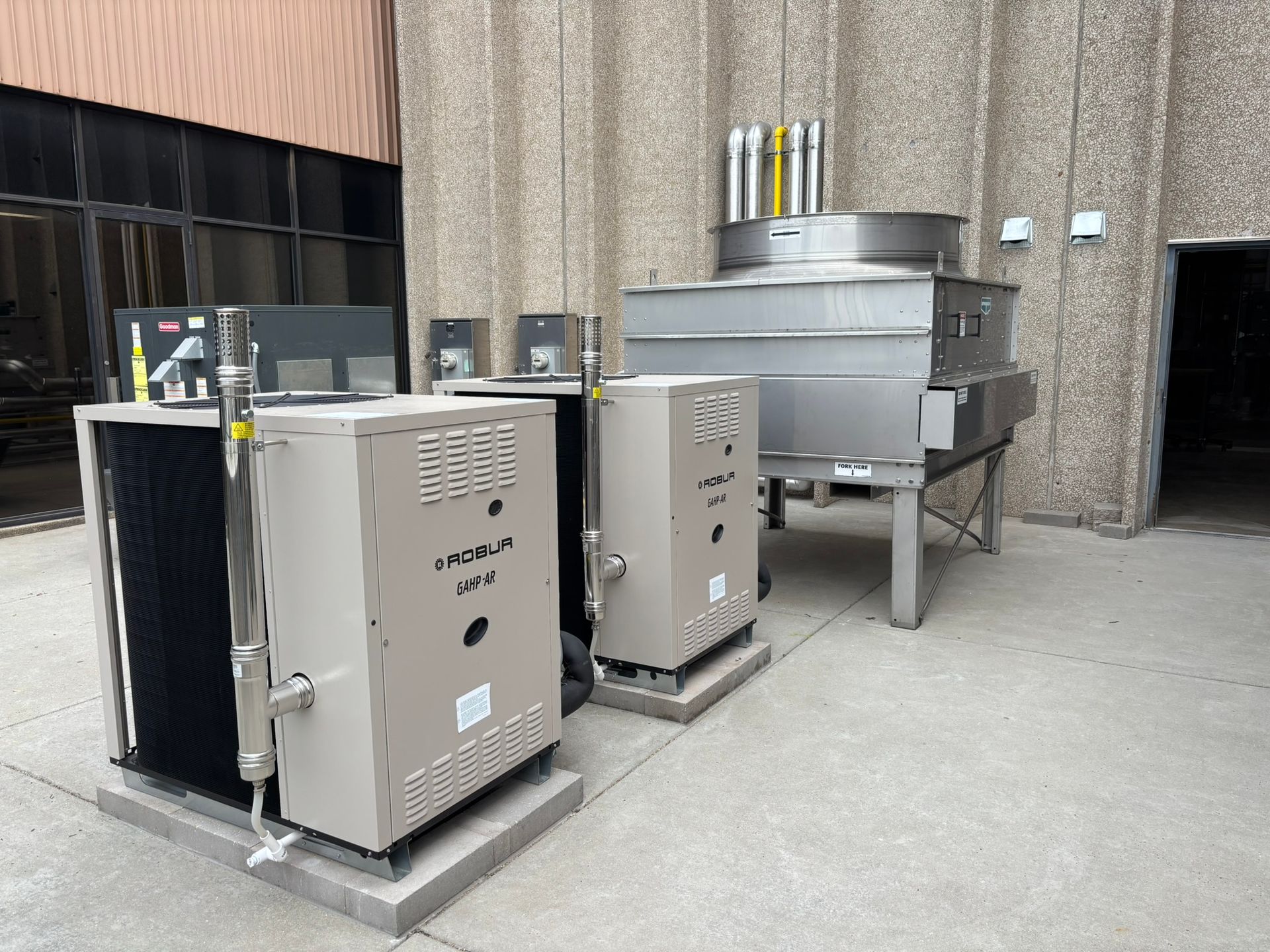 Outdoor mechanical equipment, including two beige units, a large metal cooler, and chimneys against a concrete building.