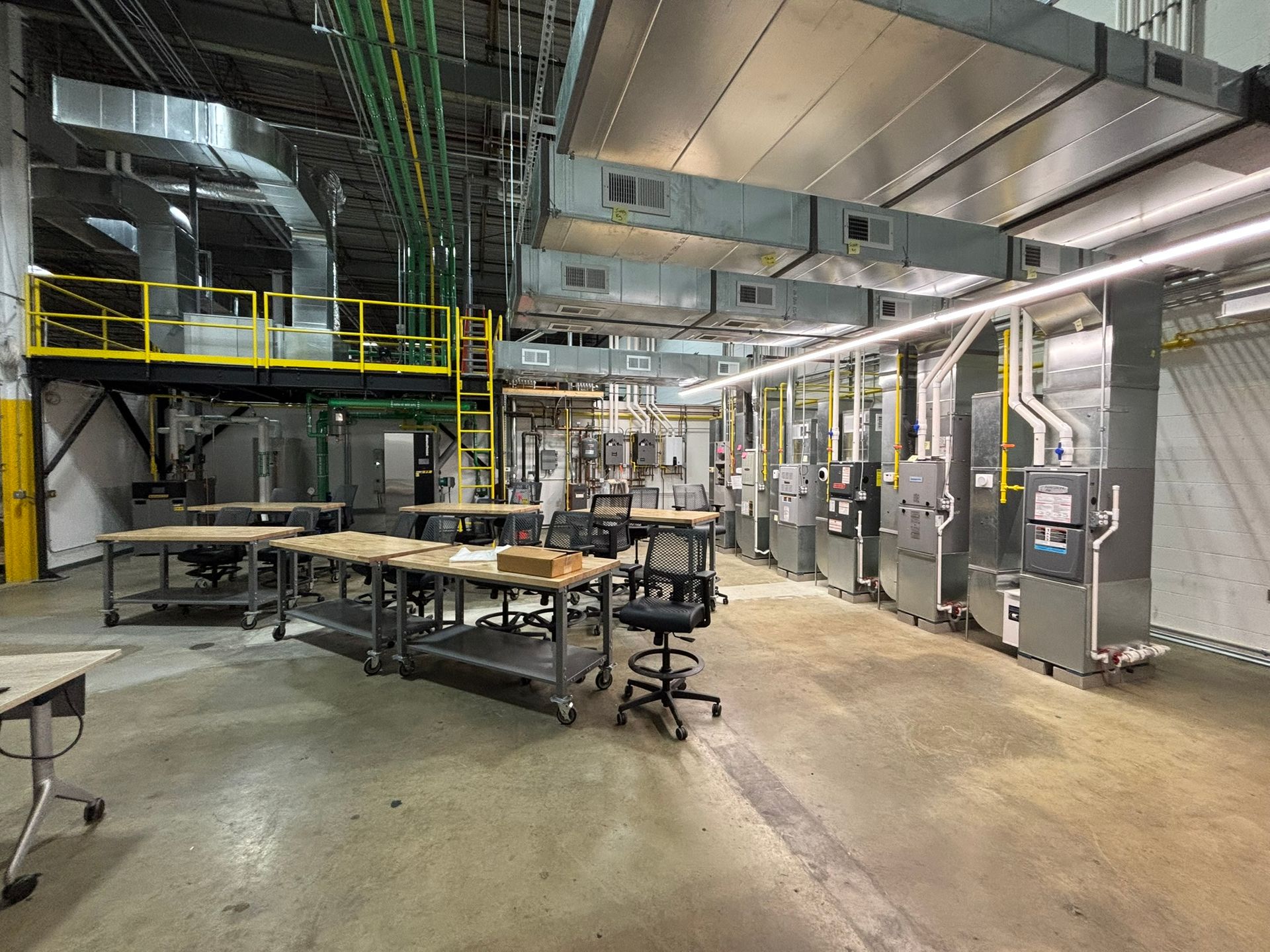Factory interior with machinery, tables, and ductwork. Overhead pipes and yellow safety rails.