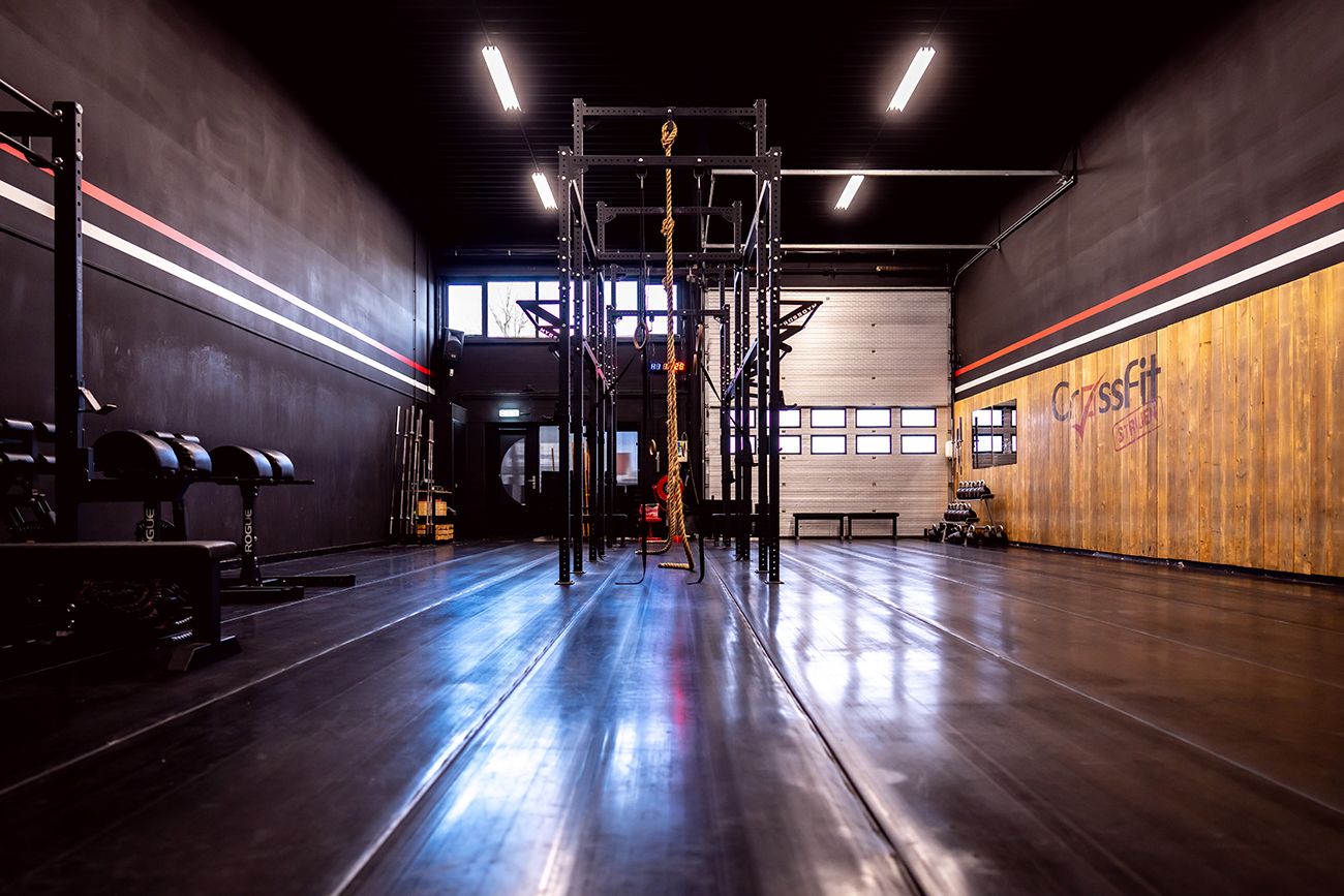 Interieur van een sportschool met trainingsapparatuur en open ruimte; donkere muren, glanzende vloer, raam aan de achterkant.