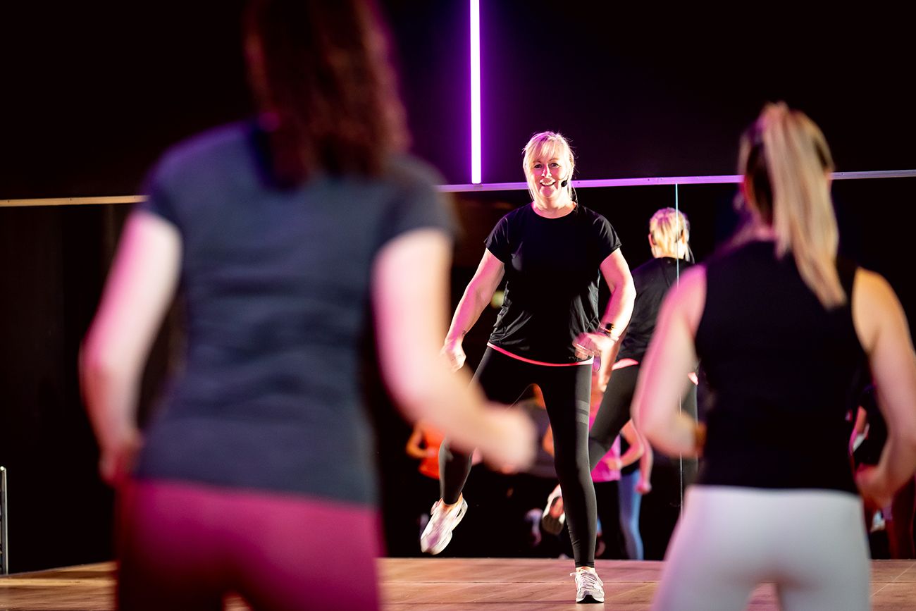 Groepsfitnessles in een dansstudio; instructeur in zwart leidt; anderen in sportkleding volgen.