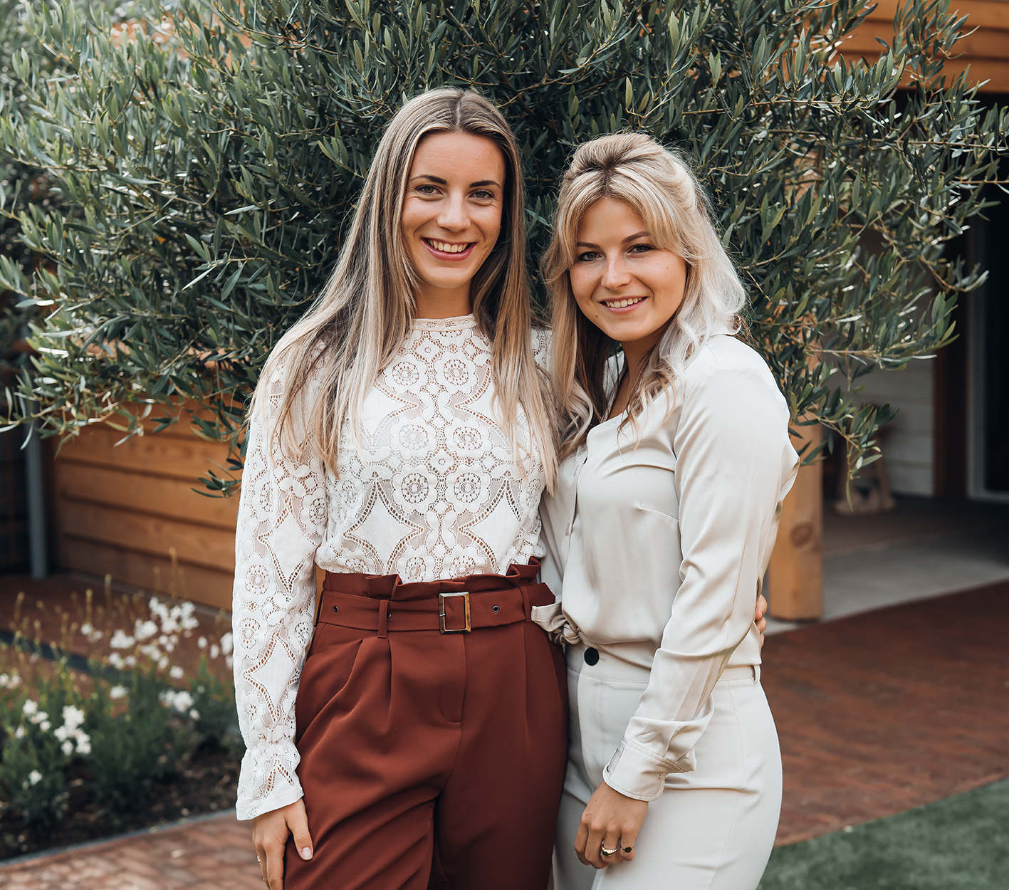 Twee lachende vrouwen poseren buiten voor het groen. De een draagt een kanten top en een roestbruine broek; de ander draagt een crèmekleurig pak.