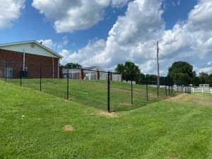 Green chain-link fence on a grassy hill; a brick building and blue sky in the background.