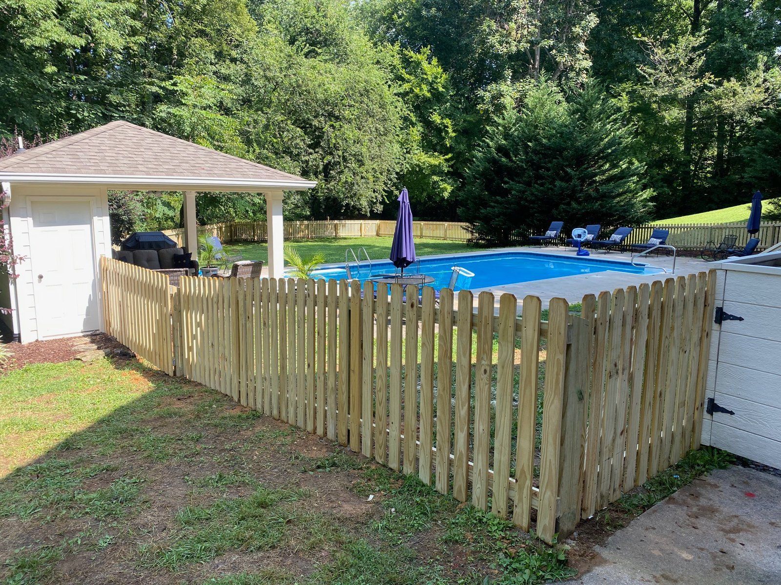 Fenced backyard with pool, gazebo, and surrounding trees. Blue pool cover, wooden fence, and open gate.