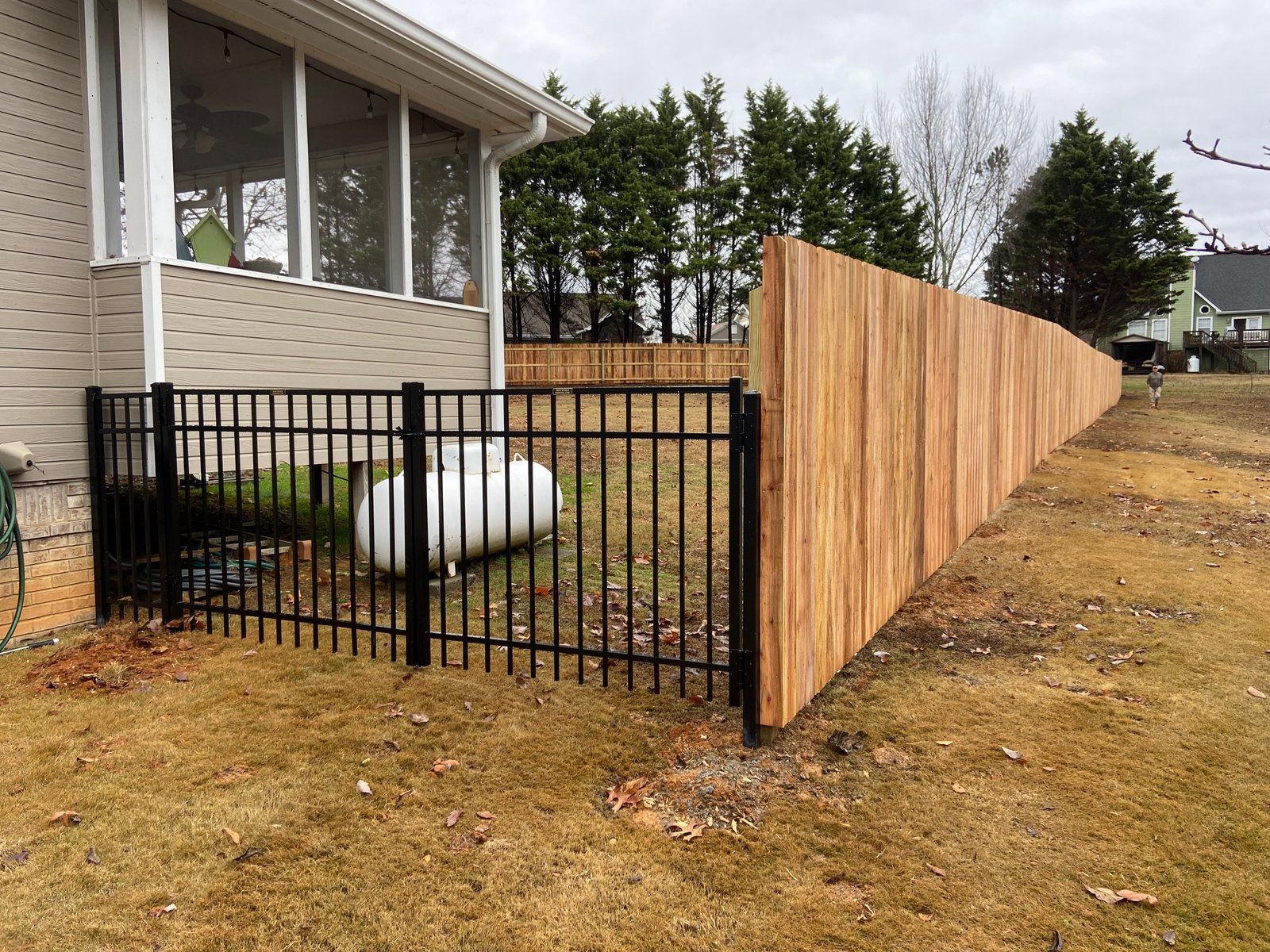 Black metal fence surrounds a propane tank next to a wooden privacy fence and house with a screened porch on a grassy lawn.