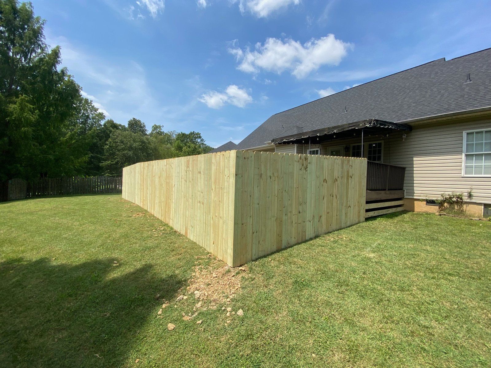 Wooden fence in a backyard, beside a house with a dark roof and green lawn under a partly cloudy sky.