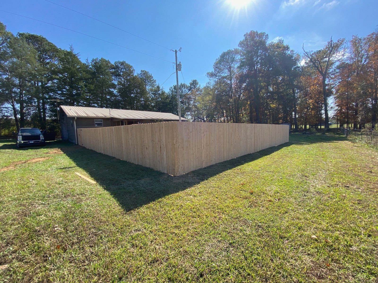 A long, wooden fence encloses a yard on a sunny day. A building and a car are in the background.