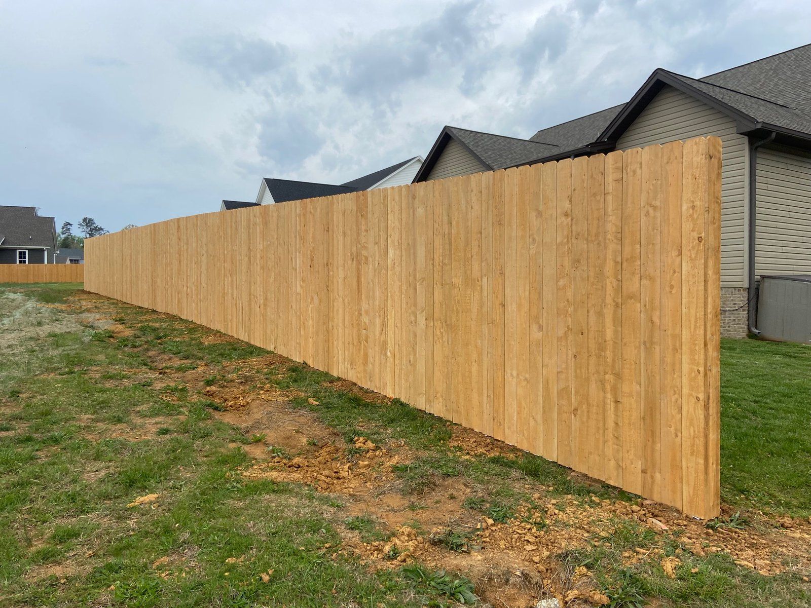 Wooden fence bordering a grassy backyard on a cloudy day, next to a residential building.