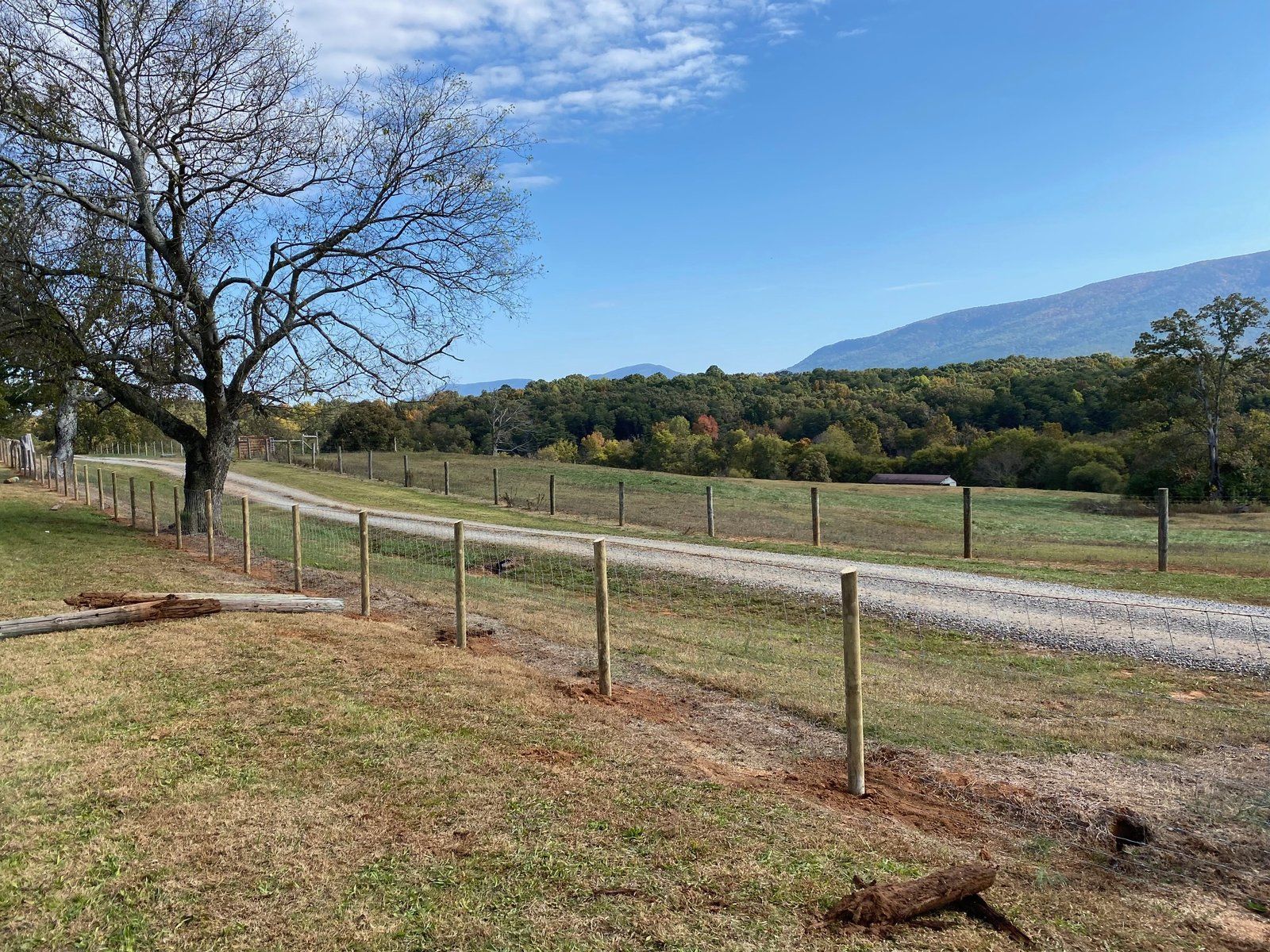 A rural scene with a fence and gravel road leading towards a mountain range under a blue sky.