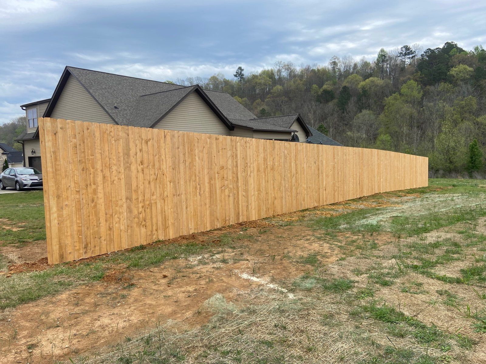 A long, new wooden fence extends across a grassy yard, with a house and trees in the background.