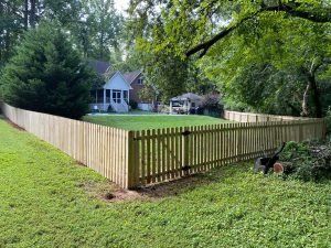 Wooden picket fence surrounding a grassy yard with a house and trees in the background.