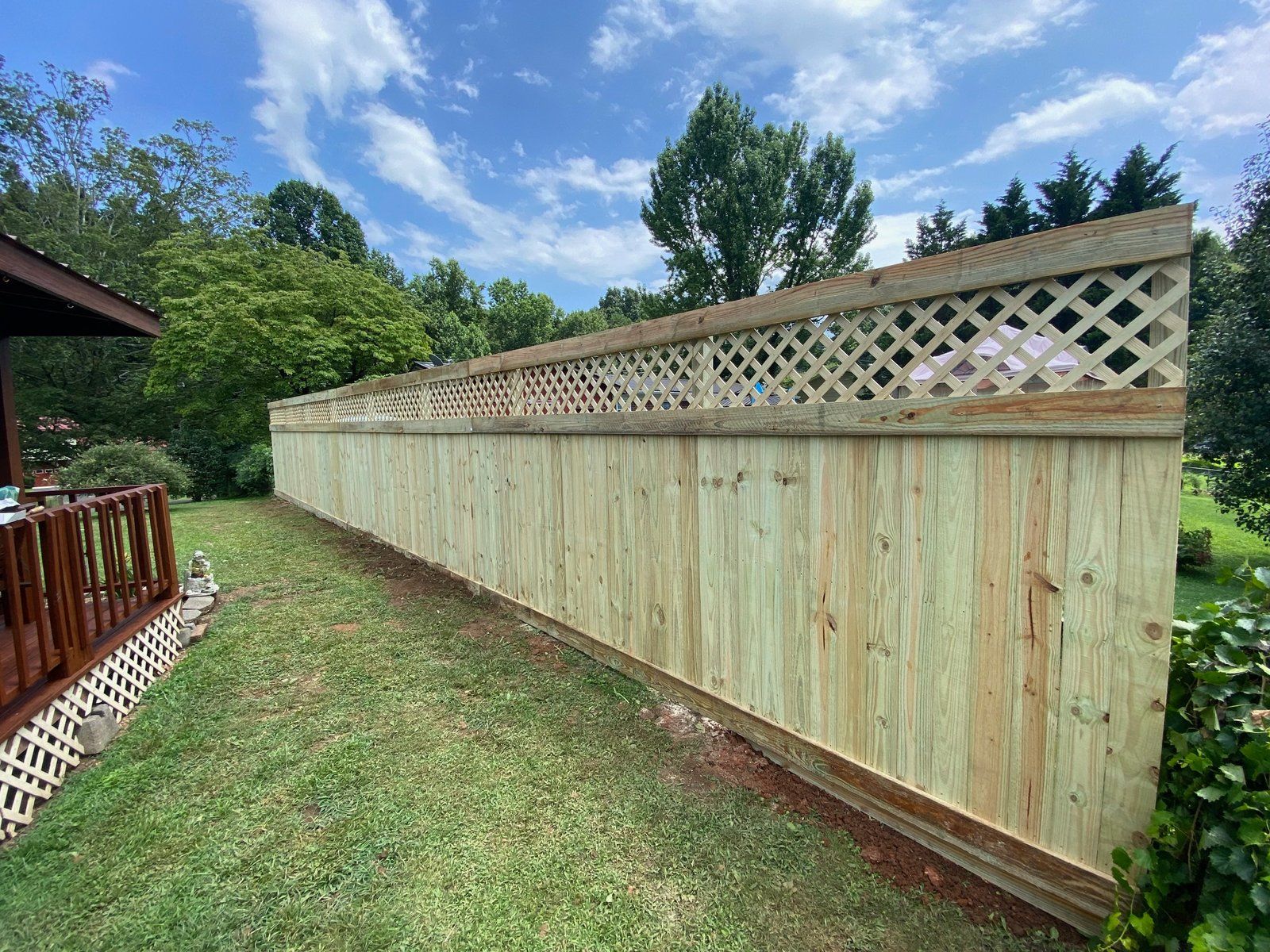 Wooden fence with lattice top in a yard, with green grass and trees, against a blue sky.