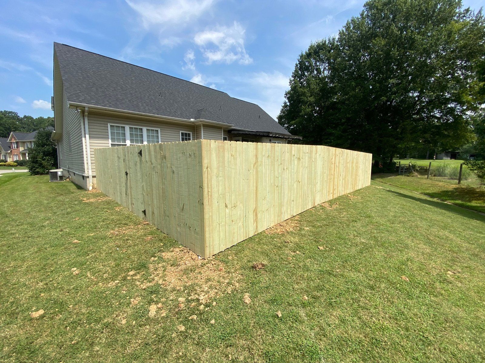 A wooden fence surrounds a house with a dark roof on a sunny day. Green grass surrounds it.