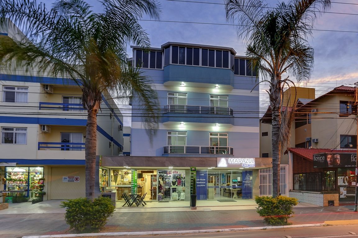 Edifício de hotel com fachada azul, lojas no nível da rua, palmeiras e céu crepuscular.
