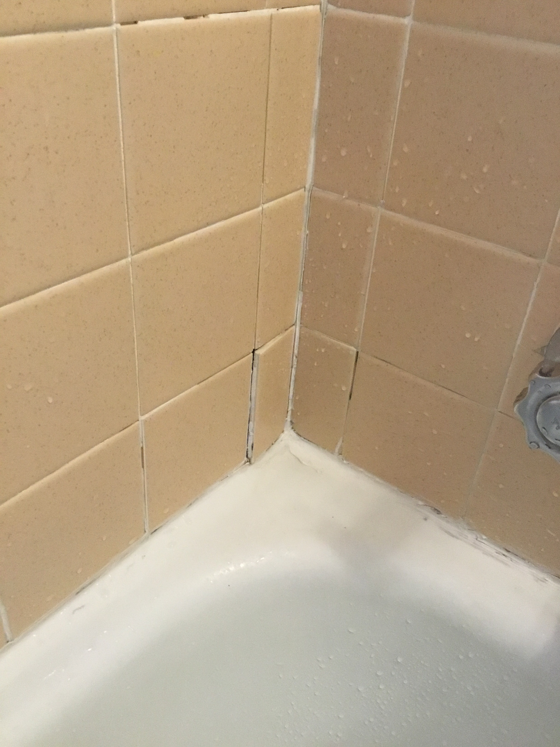 Beige tiled bathroom corner, white caulk along tub, some grout is missing.
