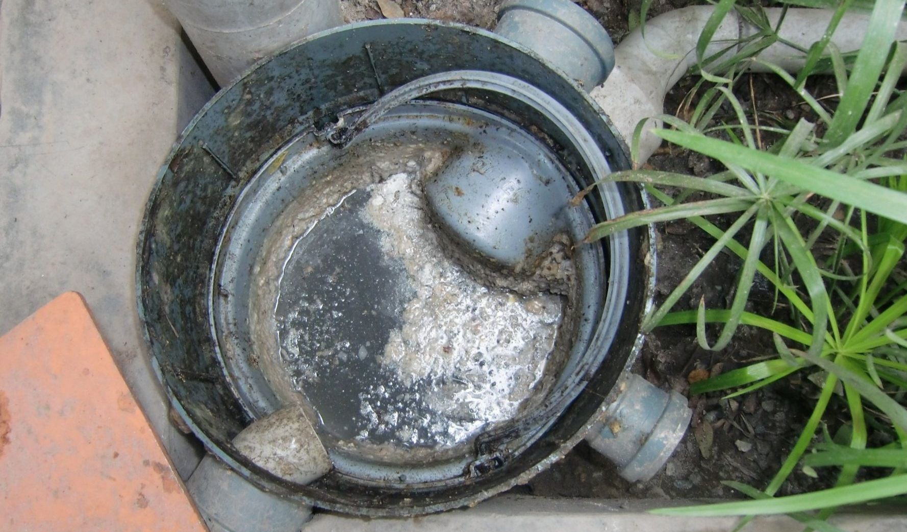 Top-down view of a circular, open grey drainage fixture with debris inside and gray pipes on its side.