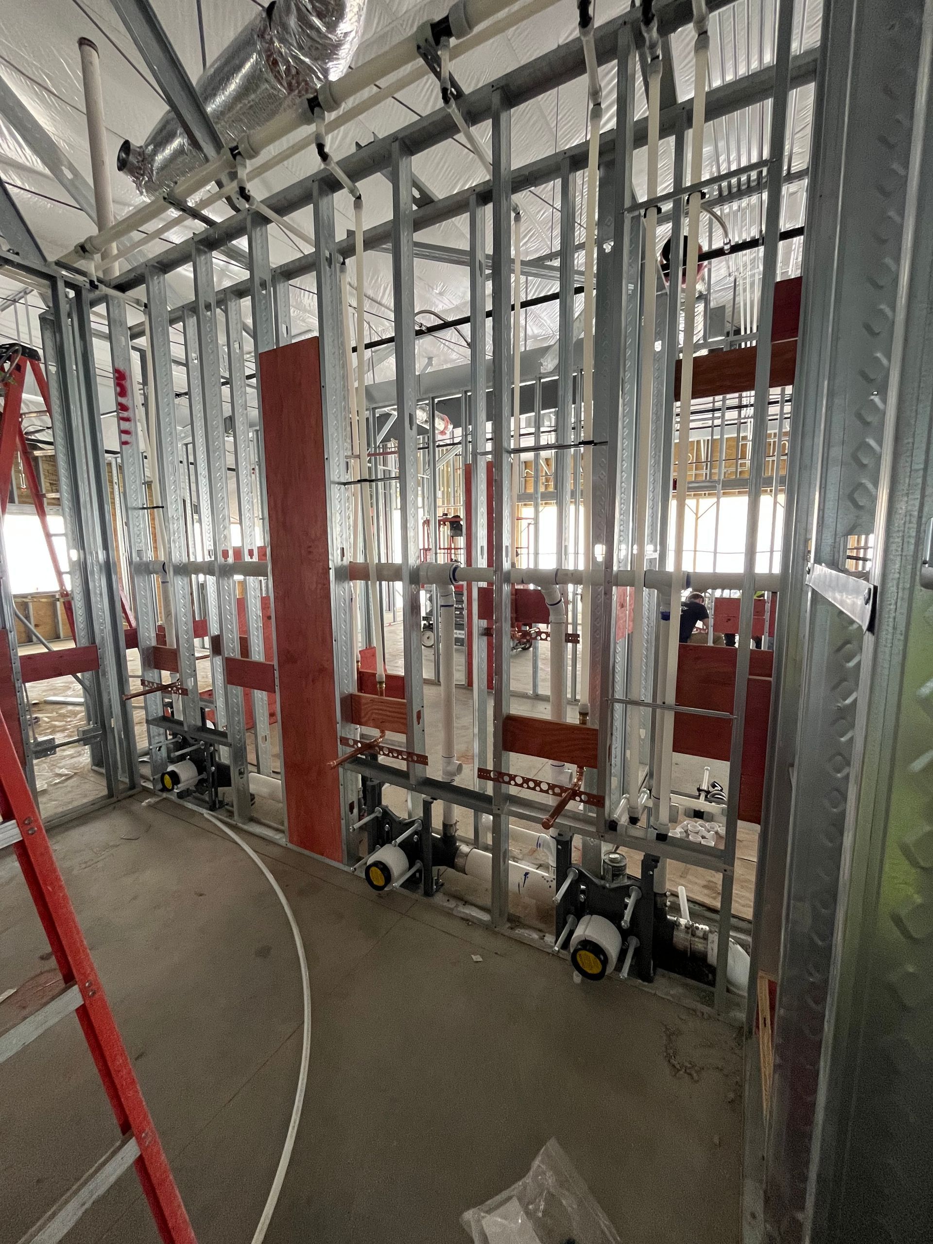 Construction site interior with metal framing, pipes, and electrical wiring. Red ladder leans on the left.