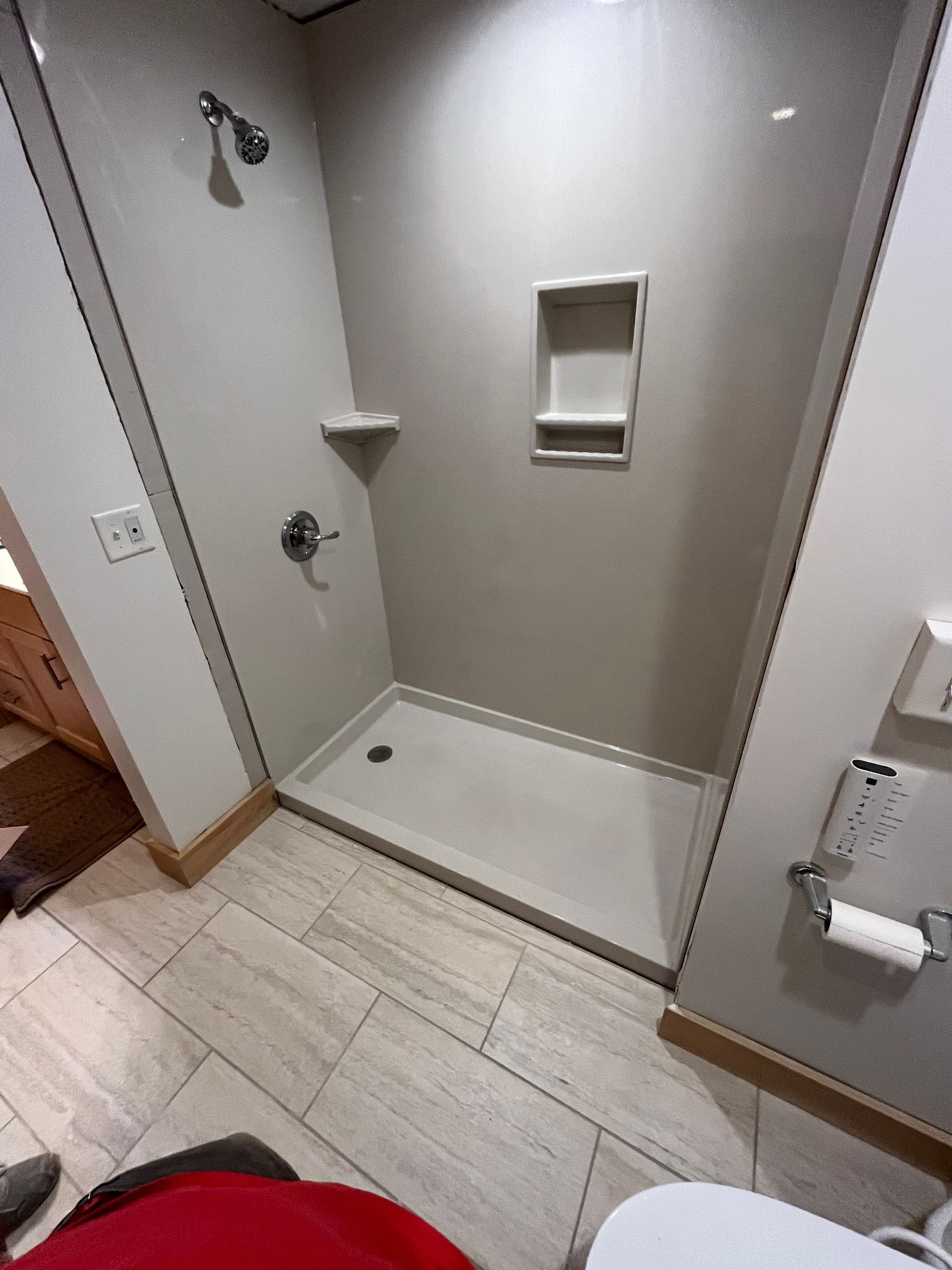Shower stall with neutral-toned walls, built-in shelf, and a showerhead. White tile flooring and a toilet are visible.