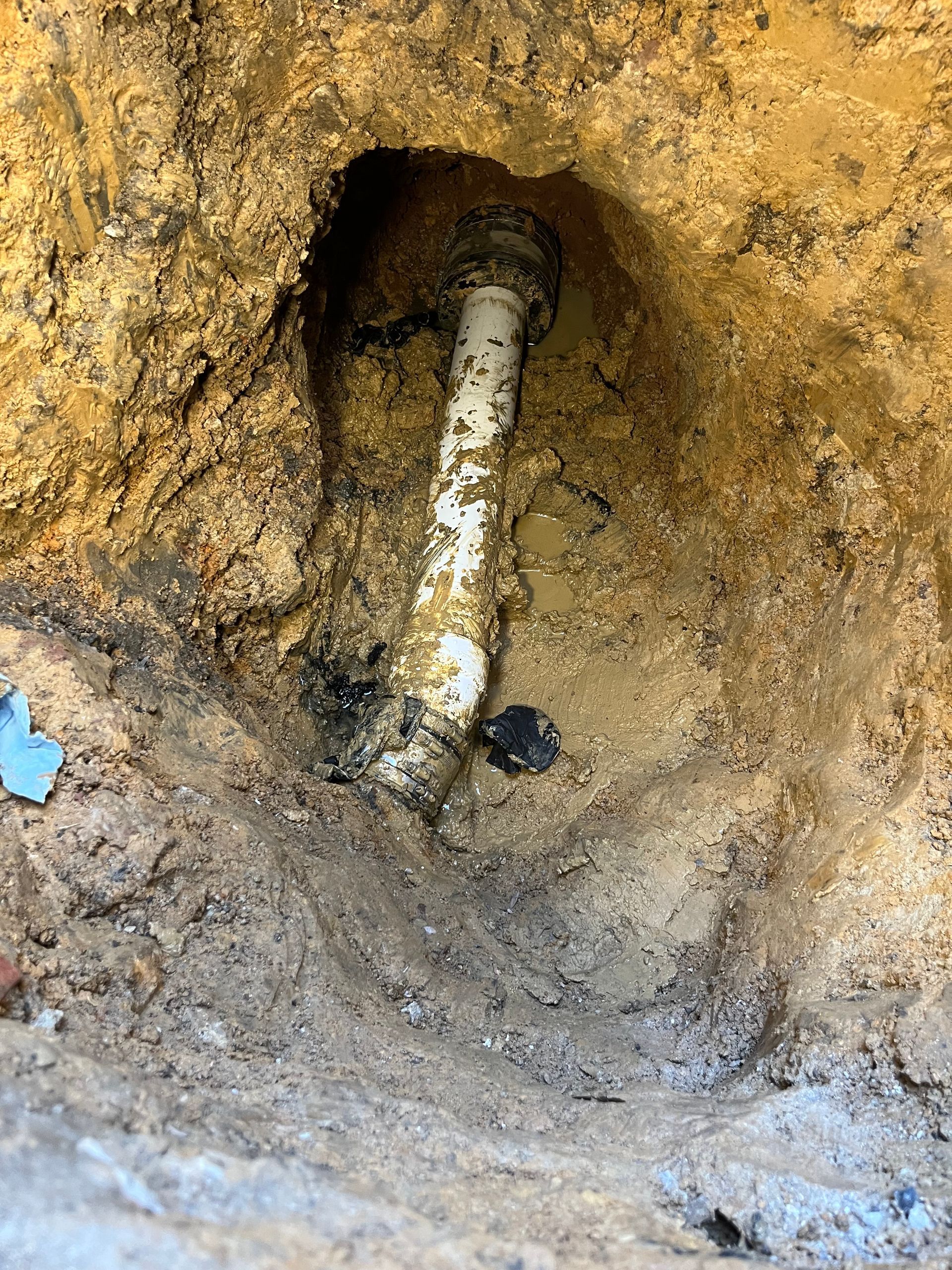 White PVC pipe exposed in a hole dug in dirt, likely a plumbing repair.
