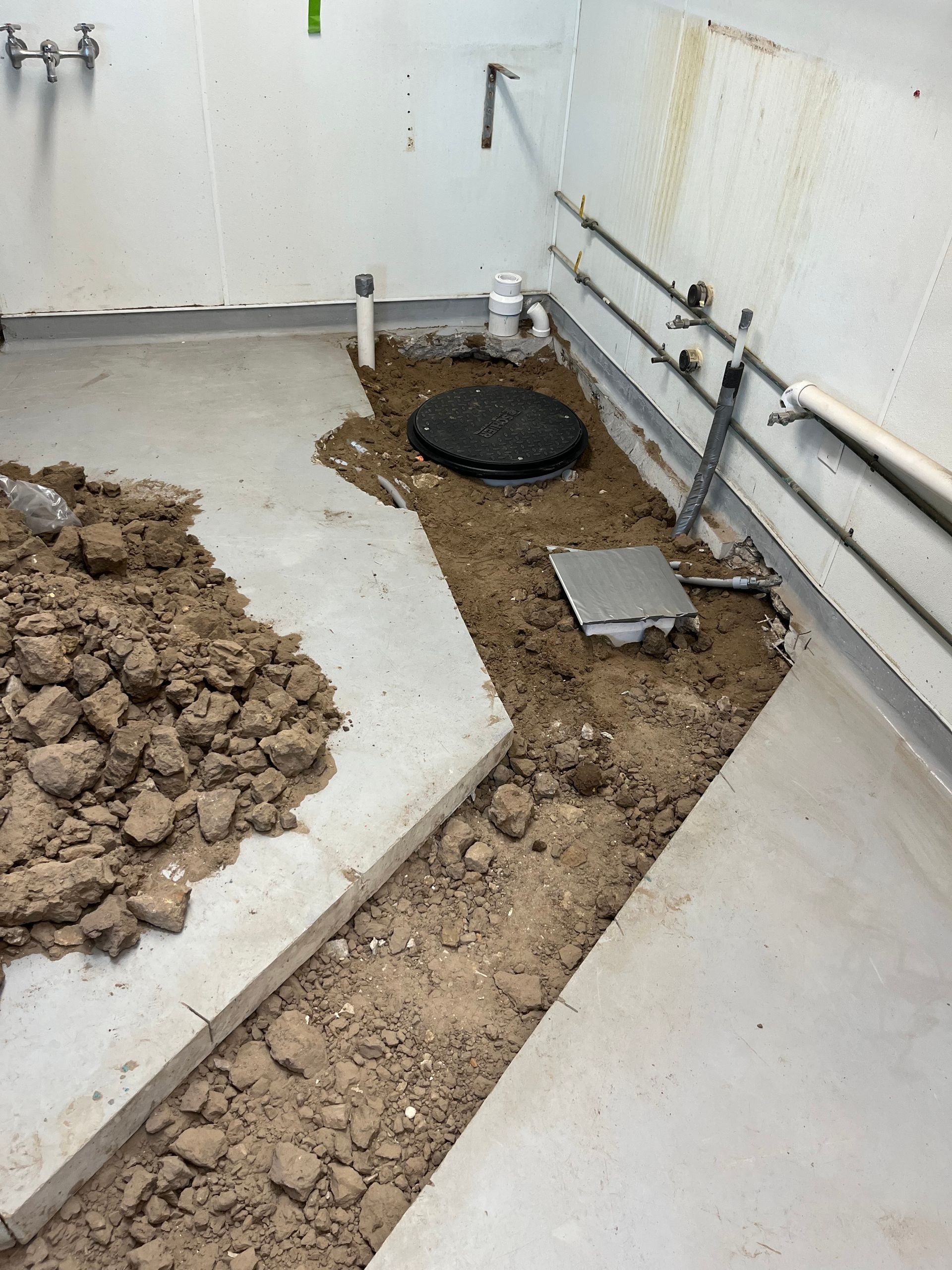 A construction site: exposed plumbing and a trench in a concrete floor, surrounded by dirt.