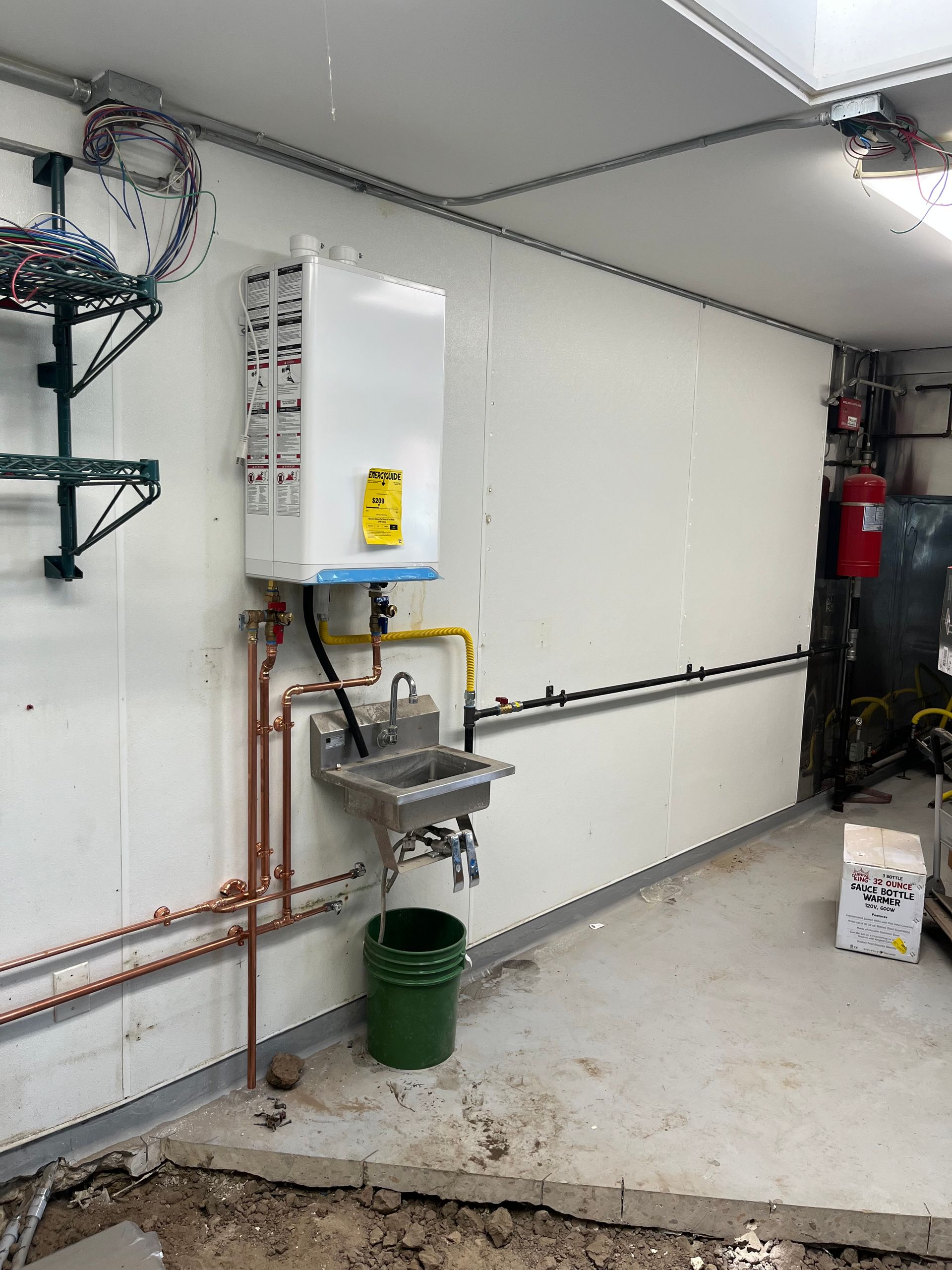 A wall-mounted water heater above a sink and a green trash can. Copper and yellow pipes run along the wall.