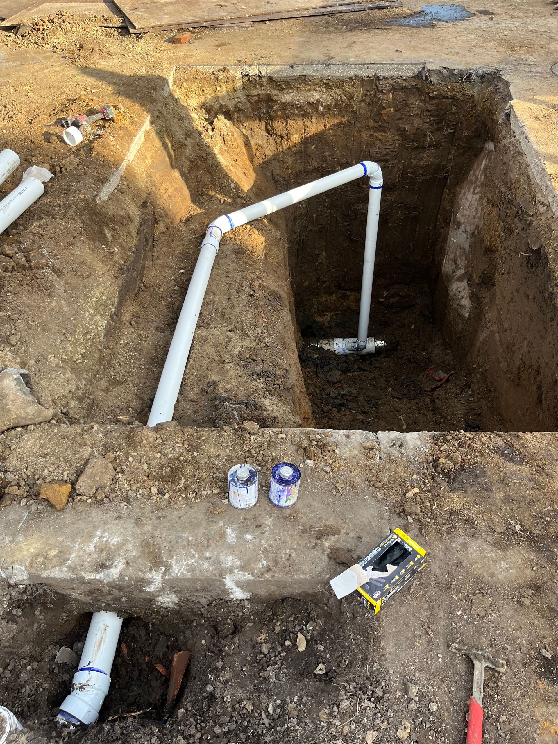 White PVC pipes installed in an excavated pit, with tools and materials nearby.