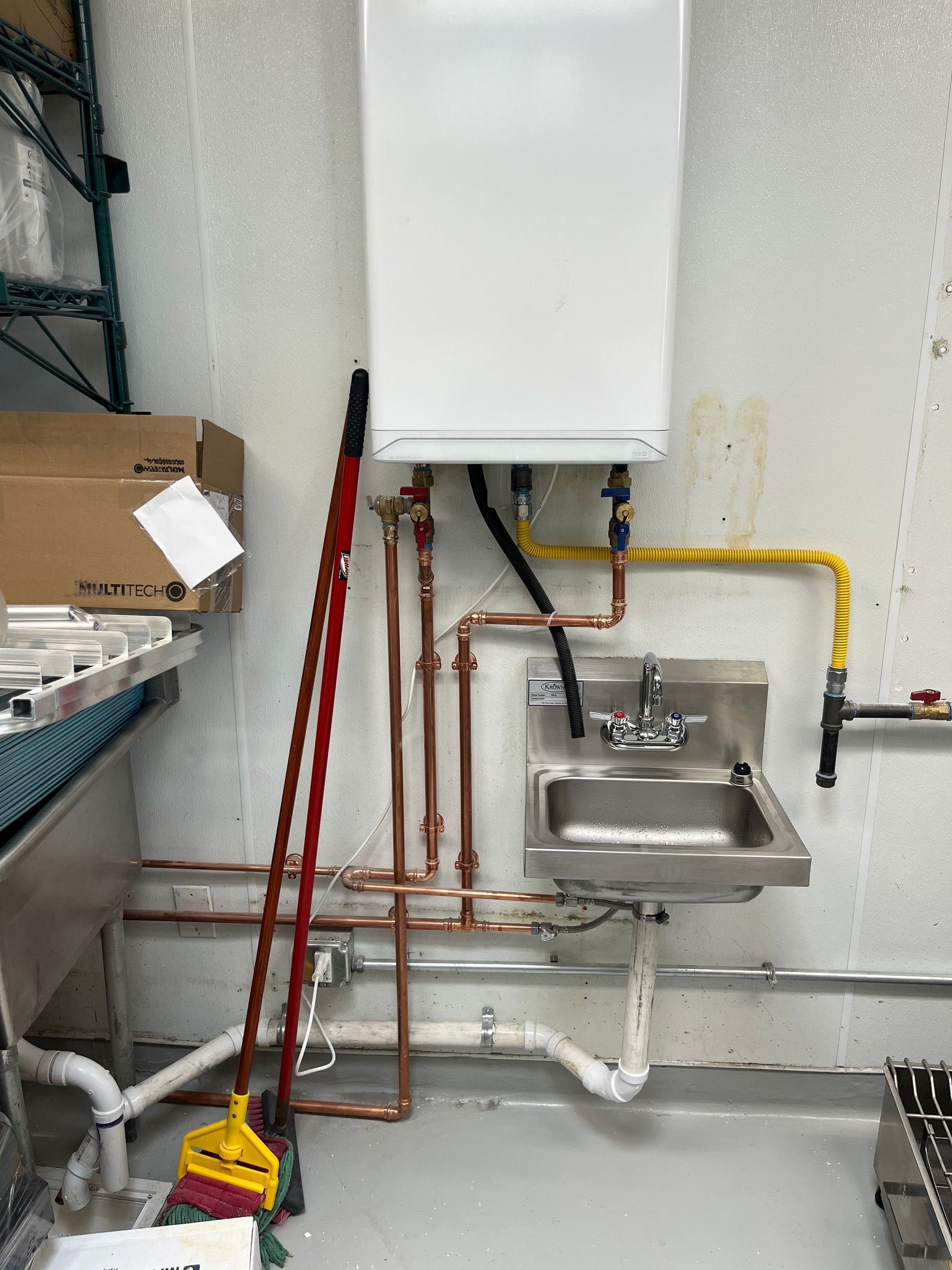 Wall-mounted water heater above a stainless steel sink, with copper pipes and a yellow gas line. A broom leans nearby.