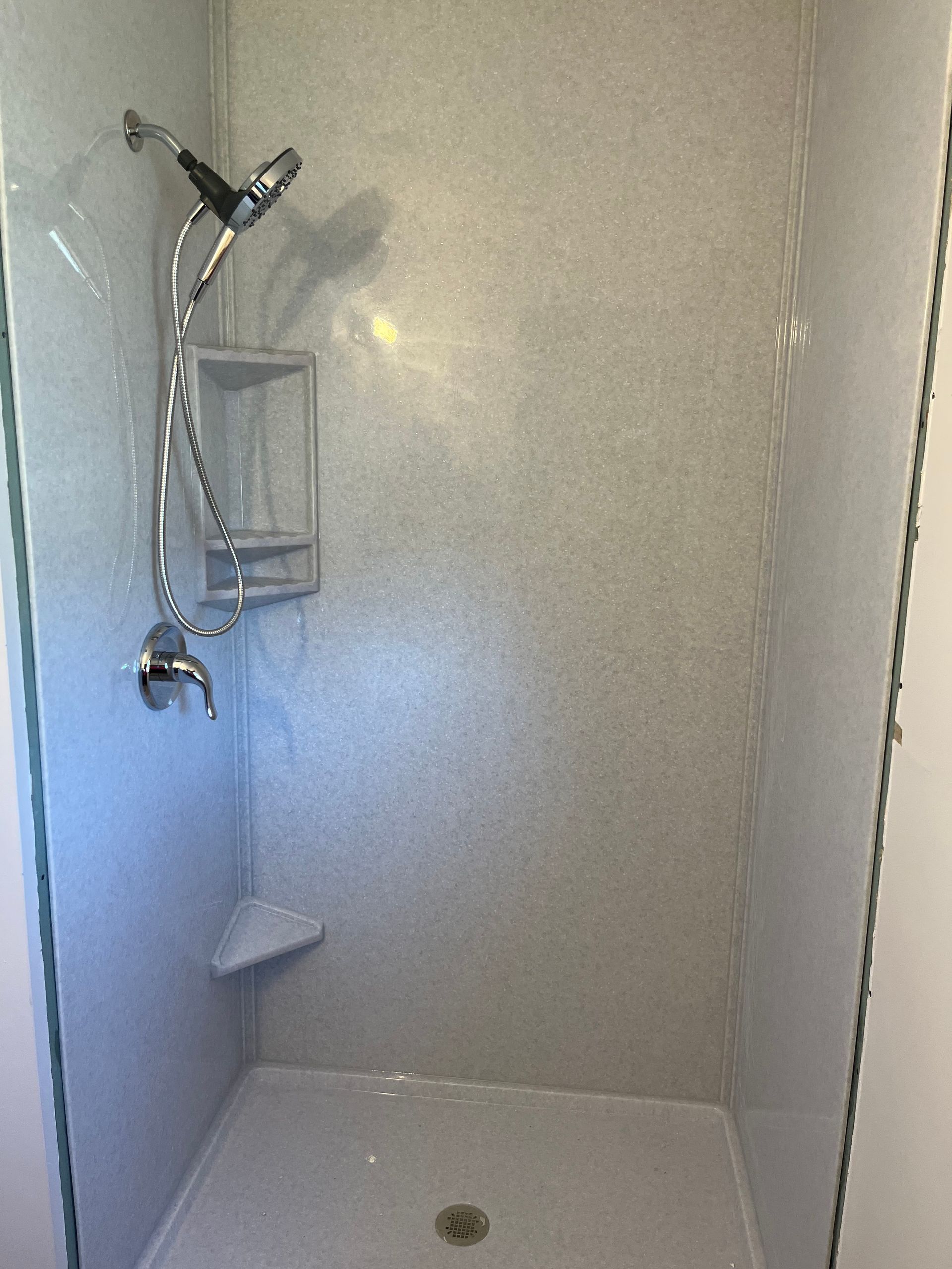 A walk-in shower with light gray, speckled walls, a handheld showerhead, chrome fixtures, and a corner shelf.