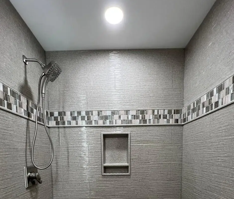 Shower with gray textured tiles, decorative band, and recessed shelf. Chrome showerhead and fixtures, bright ceiling light.