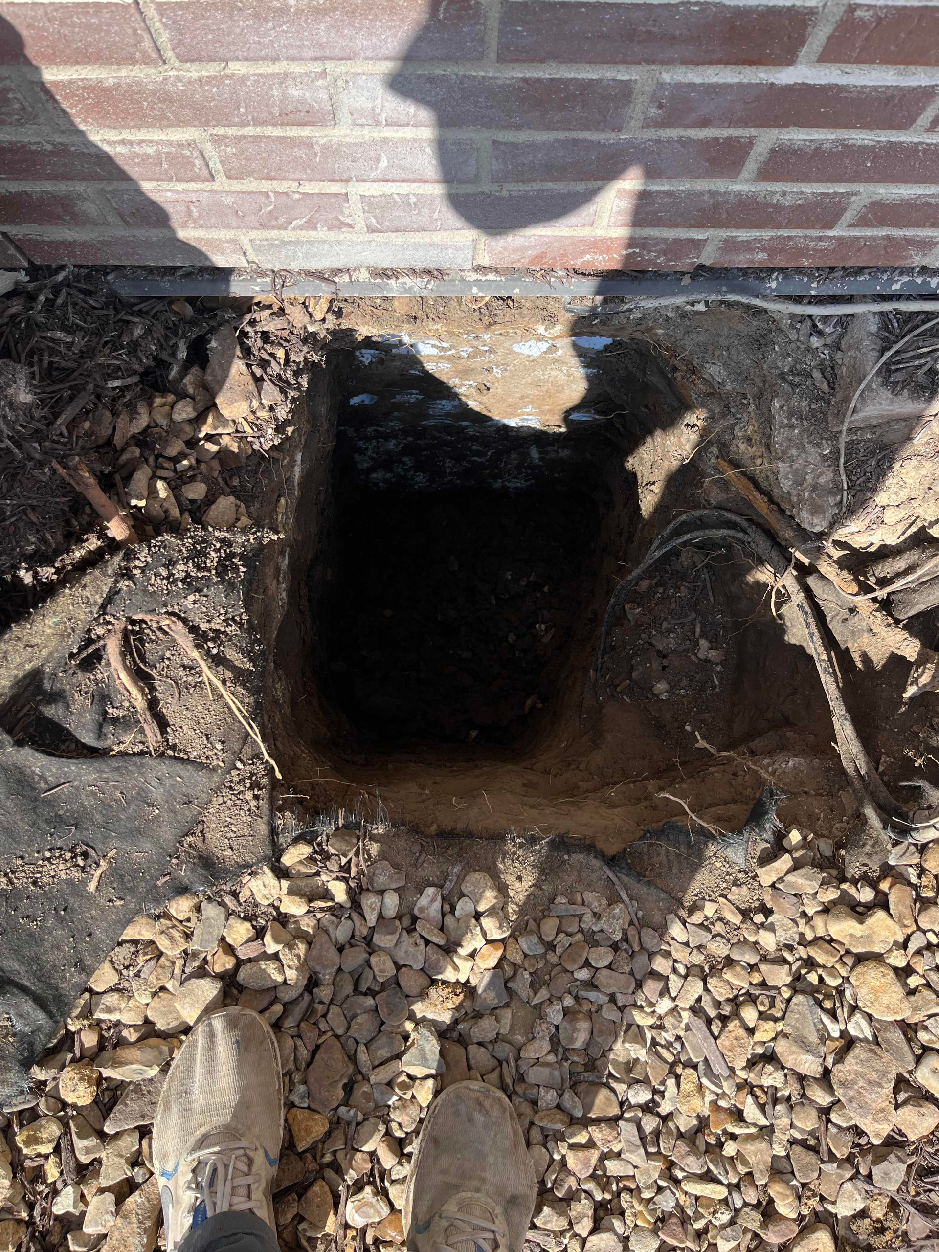 Hole dug next to a brick wall with exposed dirt and gravel, view from above.