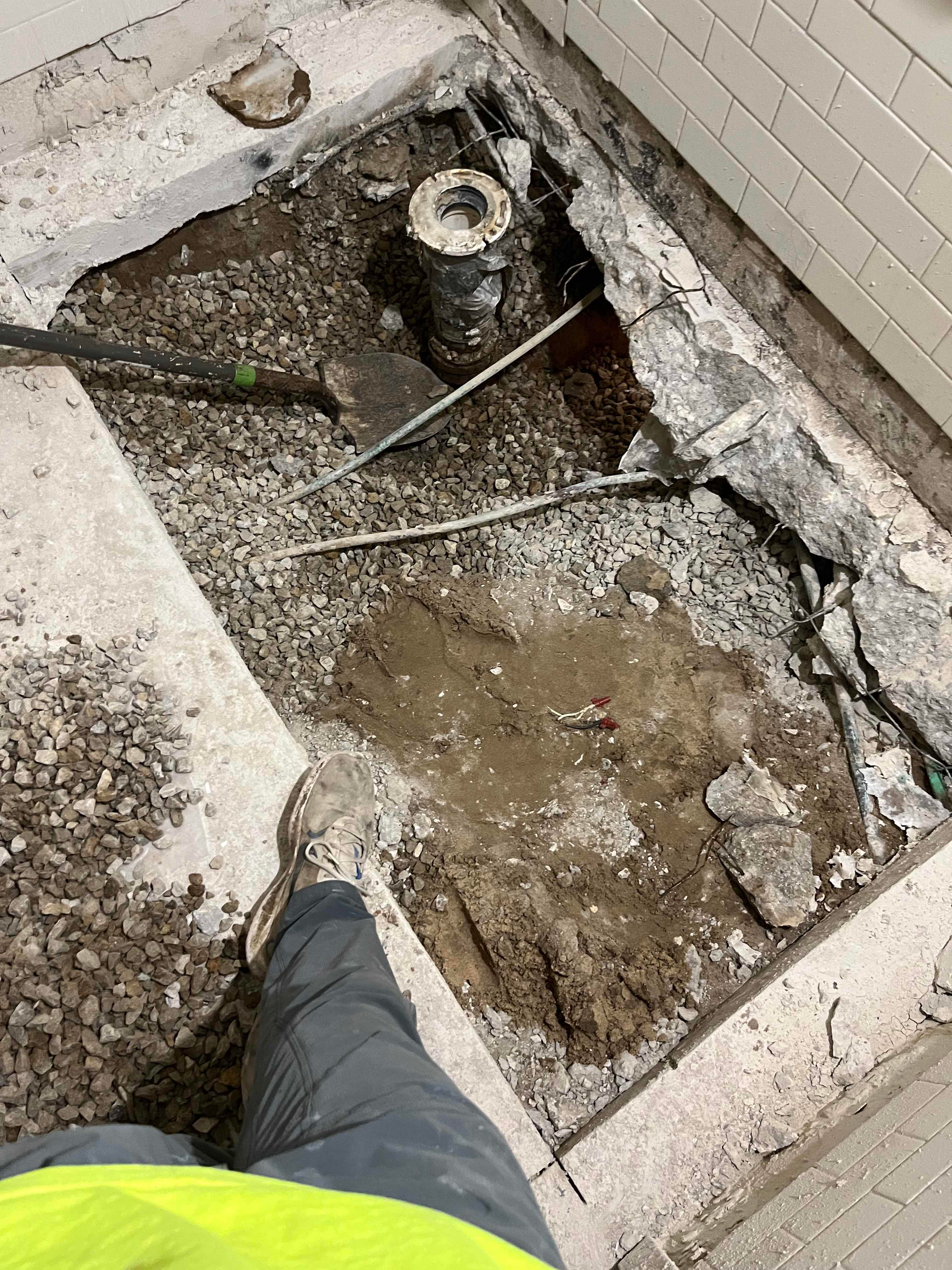 Person standing over an open hole in a concrete floor, revealing plumbing and gravel.