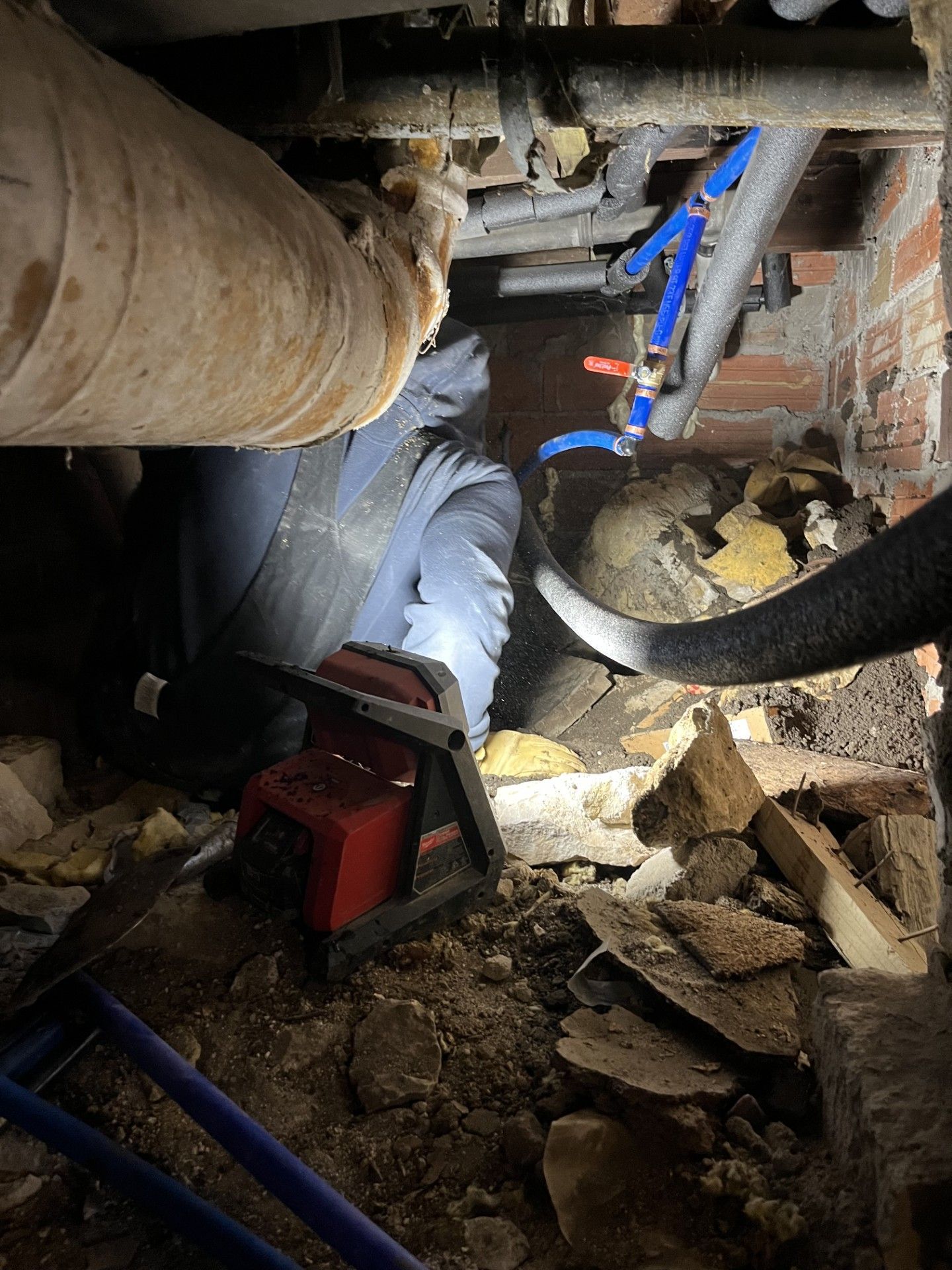 A worker kneels in a dark, dirt-floored crawlspace, performing plumbing repairs on blue PEX pipes near a brick wall.