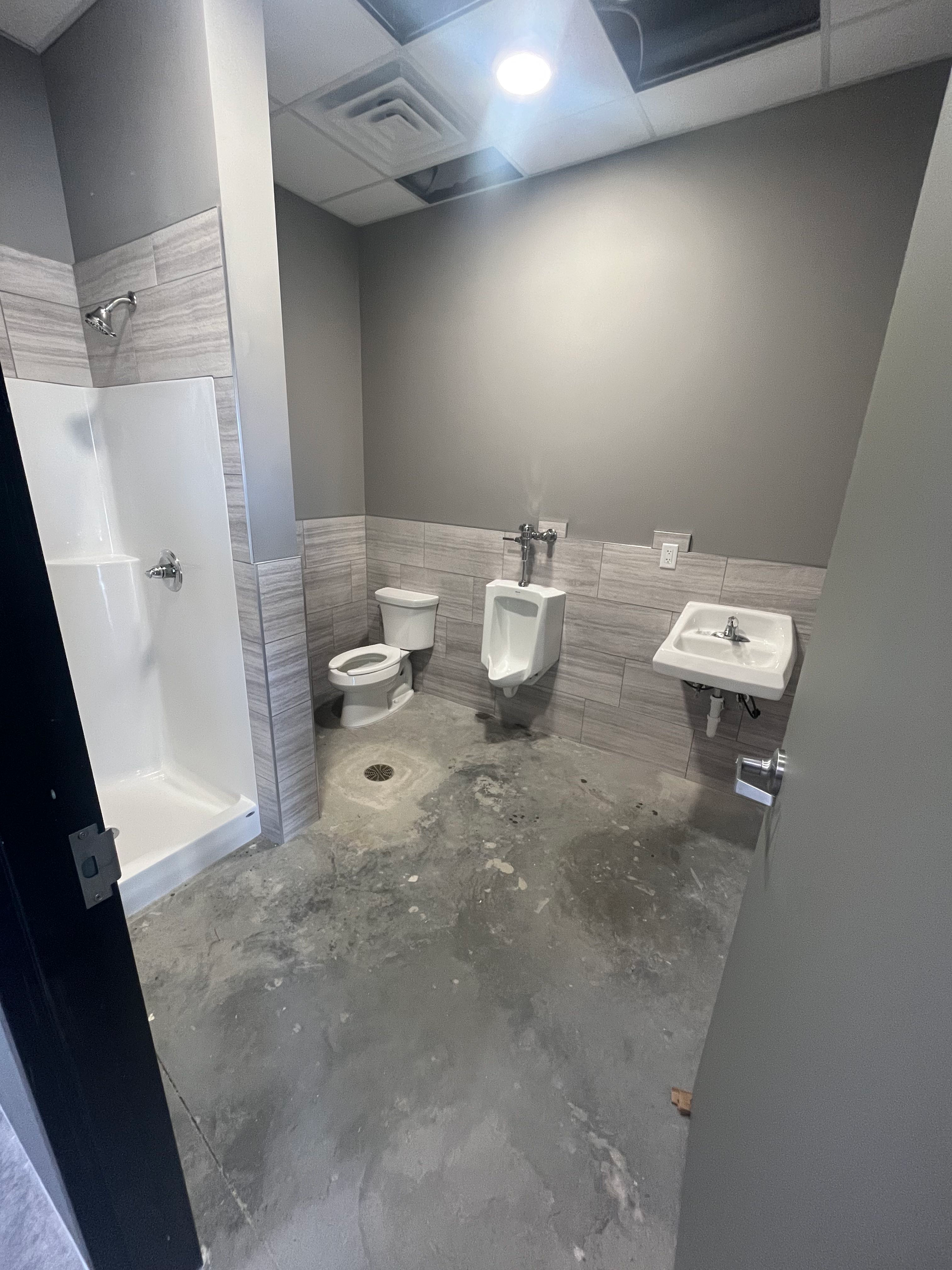 Unfinished commercial restroom with a toilet, urinal, shower, and sink; gray and white color scheme.