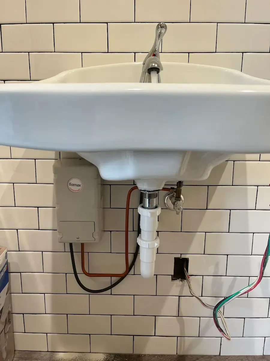 White ceramic sink with faucet against white brick tiles. An electric water heater and exposed wires are visible.