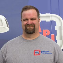 A person with a goatee wears a grey shirt with an Advanced Plumbing, Inc. logo, standing in front of a blue company van.