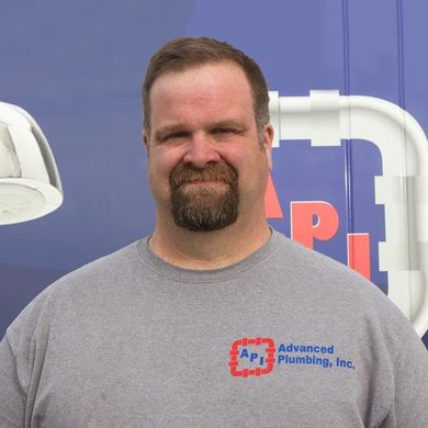 A person with a goatee wears a grey shirt with an Advanced Plumbing, Inc. logo, standing in front of a blue company van.