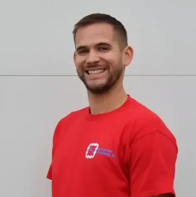 A person smiling, wearing a red t-shirt with a company logo, standing against a light gray wall.