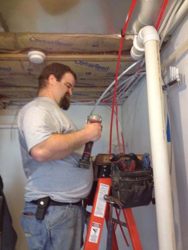 Man on ladder, using a power tool near pipes in a basement, wearing work clothes.