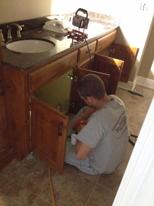 A person is working under a bathroom vanity, cabinets open, tools visible.
