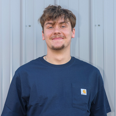 A person with messy brown hair and a goatee smiles, wearing a navy blue short-sleeved pocket t-shirt against a gray wall.