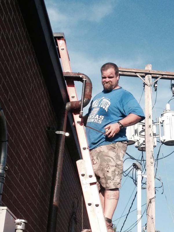 Man on ladder near brick building and power lines. He’s wearing a blue shirt and camo shorts, looking at the camera.