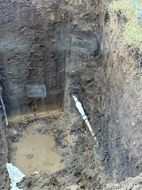 An excavation pit revealing buried water pipes. Muddy soil, a small puddle, and a blue-and-white pipe are visible.