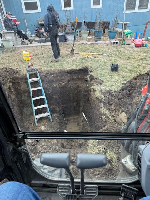 A backhoe operator's view of a large excavation in a backyard. A person stands nearby, other people are in the background.