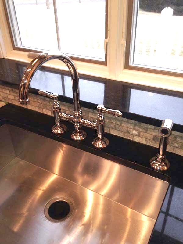 Stainless steel kitchen sink with chrome faucet and sprayer, set against black countertop and window.